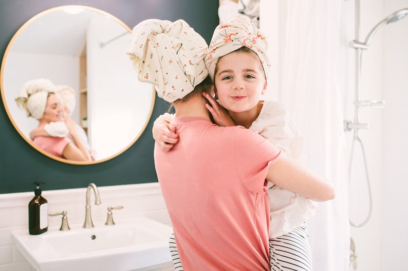 May include: A woman and a young girl are in a bathroom, both wearing white towels wrapped around their heads. The woman is wearing a pink shirt and the girl is wearing a white shirt with black and white stripes. They are smiling and hugging each other.