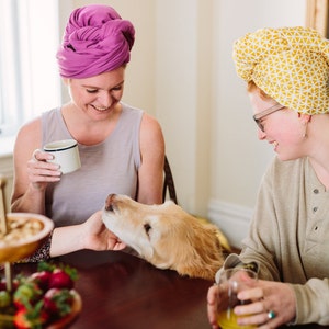 May include: Two people are sitting at a table with a golden retriever dog. The person on the left is wearing a purple turban and holding a white mug. The person on the right is wearing a yellow turban and holding a glass of orange juice. The dog is resting its head on the table.
