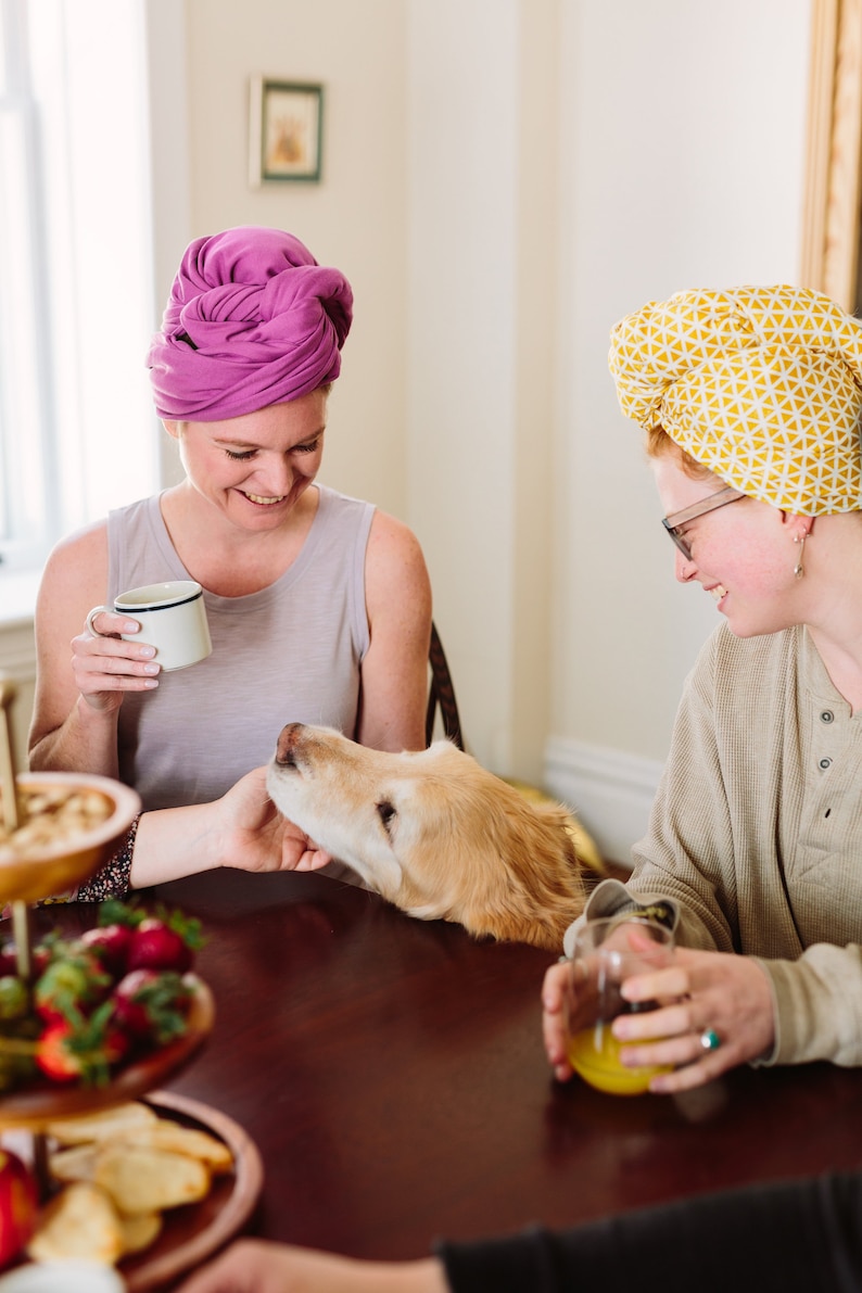 May include: Two people are sitting at a table with a golden retriever dog. The person on the left is wearing a purple turban and holding a white mug. The person on the right is wearing a yellow turban and holding a glass of orange juice. The dog is resting its head on the table.