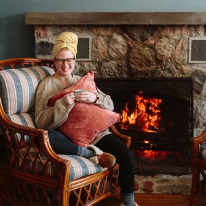 May include: A woman wearing a yellow patterned headscarf sits in a wicker chair in front of a fireplace. She is holding a mug and smiling. The fireplace is lit and has a stone surround.