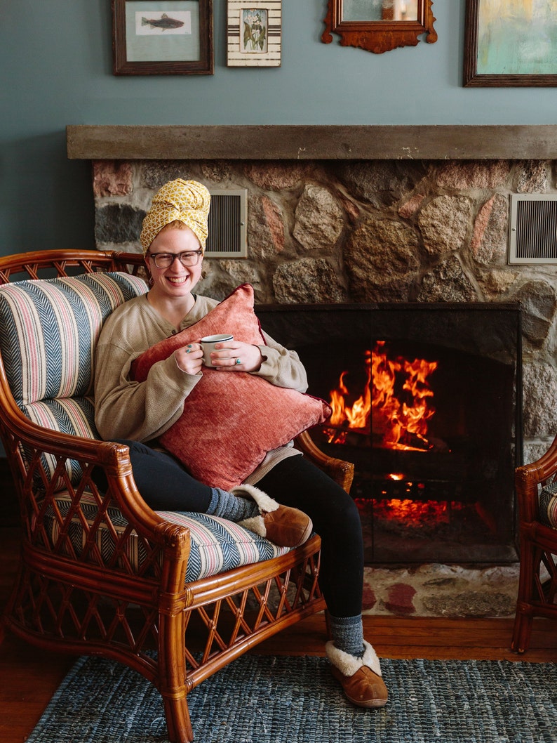May include: A woman wearing a yellow patterned headscarf sits in a wicker chair in front of a fireplace. She is holding a mug and smiling. The fireplace is lit and has a stone surround.