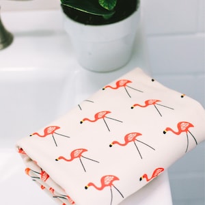May include: A white hand towel with a pink flamingo pattern. The towel is folded and sitting on a white countertop.