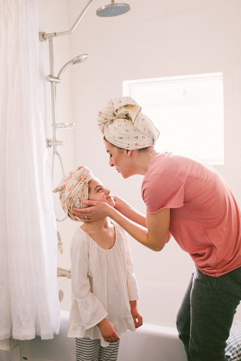 May include: A mother and daughter in a bathroom, both wearing floral patterned towels on their heads. The mother is wearing a pink shirt and grey trousers, while the daughter is wearing a white shirt and black and white striped trousers.