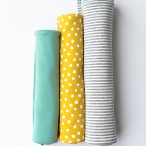 May include: Three rolled up fabric swatches. The first is a solid teal green, the second is yellow with white polka dots, and the third is grey with white stripes.