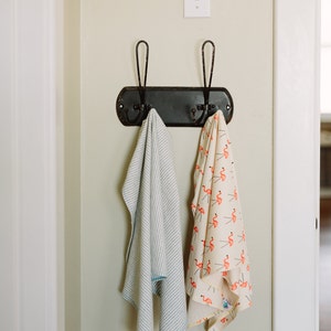 May include: Two towels hanging on a black metal hook rack with two hooks. The towel on the left is white with blue stripes. The towel on the right is white with pink flamingo illustrations.
