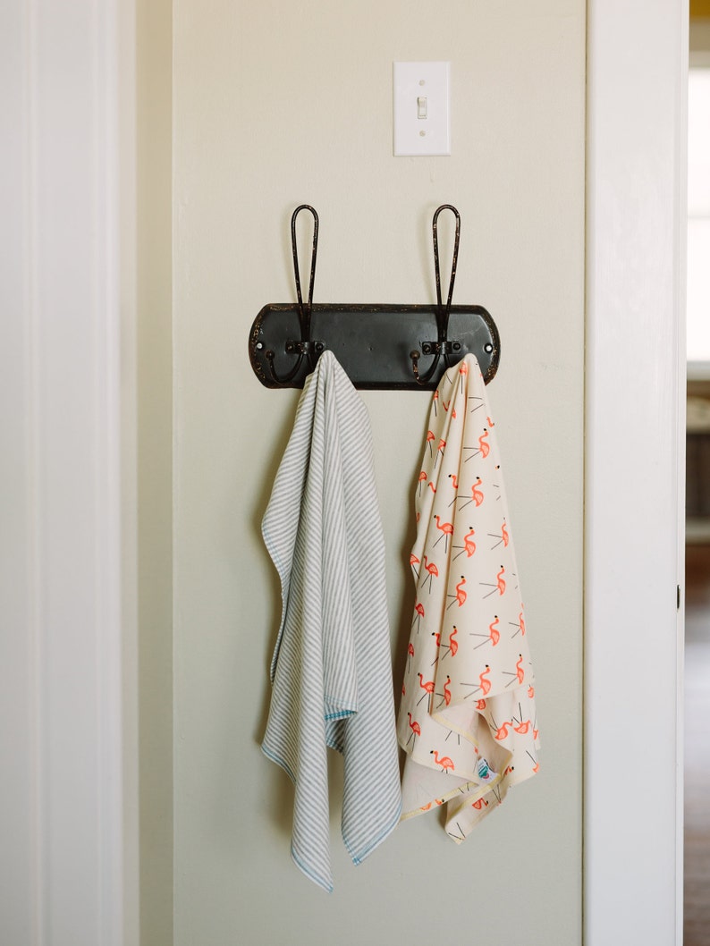 May include: Two towels hanging on a black metal hook rack with two hooks. The towel on the left is white with blue stripes. The towel on the right is white with pink flamingo illustrations.