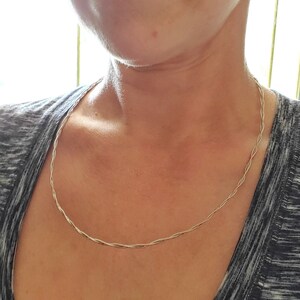 Peut inclure: Un collier en argent avec un design tressé. Le collier est porté par une personne portant un chemisier gris.
