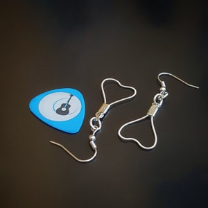 May include: A pair of silver heart-shaped earrings with a guitar pick charm. The guitar pick is blue with a black silhouette of a guitar.