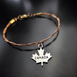 May include: A brown braided wire bracelet with a silver maple leaf charm that says "CANADA".