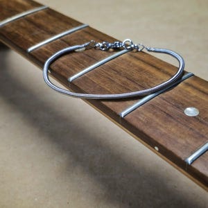 Peut inclure: Un bracelet argenté fait d'une corde de guitare, posé sur une touche de bois. Le bracelet a un design simple et élégant avec une fermeture à fermoir. La touche est marron avec des frettes argentées et de petits incrustations rondes.