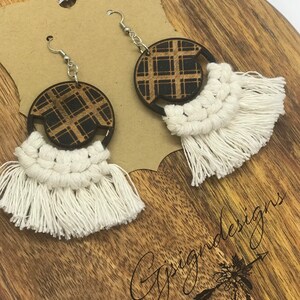 May include: A pair of handmade earrings featuring a circular wooden design with a plaid pattern. The earrings have a white macrame fringe and silver-colored ear wires. The earrings are displayed on a wooden surface.