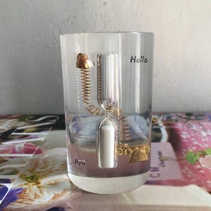 May include: Clear glass hourglass with gold-colored accents, including a telephone and a spring. The words "Hello" and "Bye" are printed on the glass. The hourglass contains white sand.