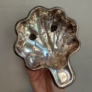 May include: A silver-toned metal dish shaped like a scallop shell with a scalloped edge and a smooth, reflective surface. The dish features a small, rounded handle extension at the bottom and two small circular indentations near the top rim.