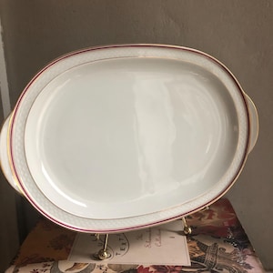 May include: Oval white serving platter with a gold rim and a thin red accent line. The platter has gold handles and is displayed on a gold metal stand. The platter is part of a tableware collection.