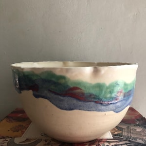 May include: A ceramic bowl with an off-white base and a wavy, hand-painted rim featuring horizontal bands of green, blue, and dark red glaze. The bowl has an irregular, organic shape with a smooth, matte finish on the lower exterior surface.