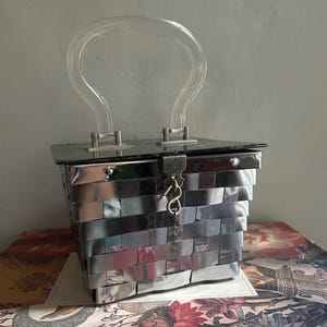 May include: A square-shaped handbag constructed from woven metallic silver strips. The bag features a clear, arched acrylic handle attached with silver-toned hardware and a hinged lid secured by a central metal latch with a small hanging hook.