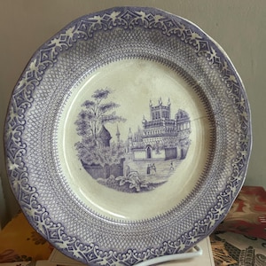 May include: A vintage ceramic plate with a blue and white design. The plate features a detailed landscape scene in the center, surrounded by a patterned border. The plate is displayed on a white stand.