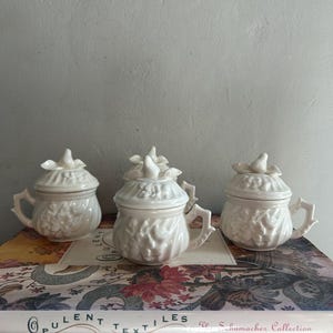 May include: Three white ceramic lidded jars with handles, each adorned with floral relief patterns. The lids are topped with a decorative finial. The jars are arranged on a patterned surface, suggesting a decorative or collectible theme.