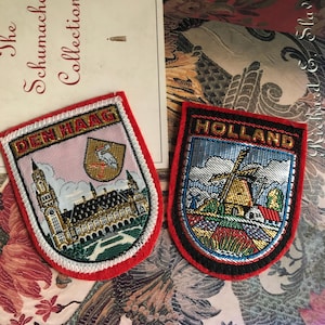 May include: Two embroidered patches. One reads "DEN HAAG" with a building and bird design, red border. The other says "HOLLAND" with a windmill scene, black border. Both are shield-shaped and colorful.