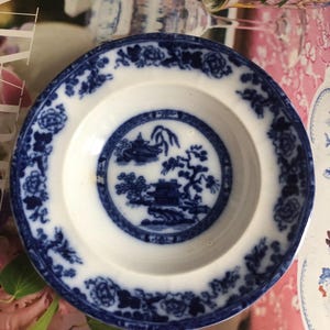 May include: A white ceramic bowl with a blue and white floral pattern. The bowl has a raised rim and a center design of a blue and white pagoda with a tree and a bird.