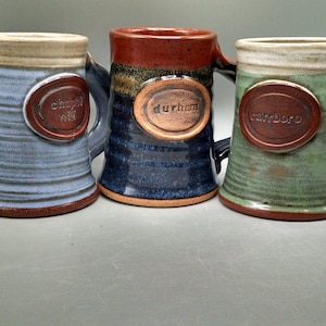 May include: Three ceramic mugs with different colored glazes. The mugs have a brown rim and a brown oval label with the words "chapel hill", "durham", and "carrboro" on each mug.