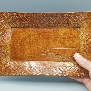 May include: A rectangular ceramic serving tray with a brown glaze and a leaf pattern around the edge. The tray features an outline of the state of North Carolina.