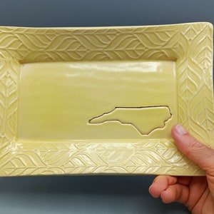 May include: A yellow ceramic serving tray with a raised leaf design border. The tray features a cutout of the state of North Carolina.