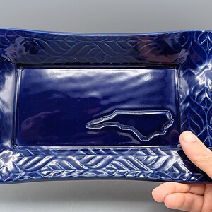 May include: A blue ceramic tray with a raised outline of the state of North Carolina. The tray has a decorative border with a woven pattern.