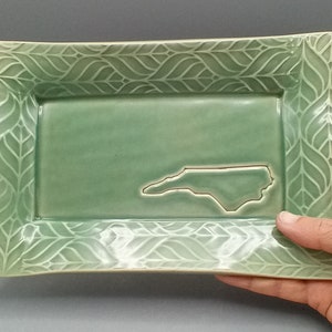 May include: A green ceramic serving tray with a raised leaf pattern around the edge. The tray features a cutout of the state of North Carolina in the center.