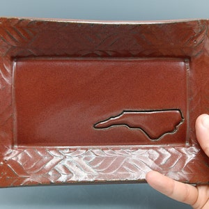 May include: Rectangular ceramic tray in a reddish-brown hue, featuring an embossed leaf pattern around the edges. The center showcases a cutout of the North Carolina state outline. A handmade decorative piece.