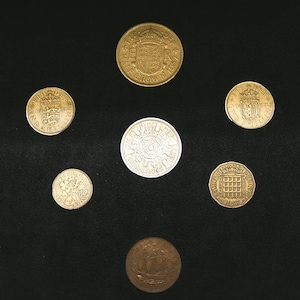 Puede incluir: Una colección de siete monedas británicas, que incluyen una media corona, un chelín, un sixpence, un threepenny bit, un two pence, un penique y un medio penique. Las monedas están dispuestas en círculo sobre un fondo negro.