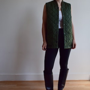 May include: A dark green quilted vest with a button closure and two pockets. The vest is worn over a white shirt and black pants.