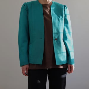 Puede incluir: Blazer turquesa con cierre de un solo botón y hombreras sutiles. La chaqueta tiene un corte entallado y un diseño clásico. El modelo lleva una camiseta marrón y pantalones negros. El blazer es de color turquesa vibrante.