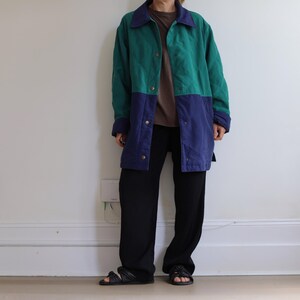 May include: A color-blocked jacket featuring a teal upper section and a navy blue lower section, paired with a brown t-shirt, black trousers, and black sandals. The jacket has a pointed collar, long sleeves, and a front button closure with metal snaps.