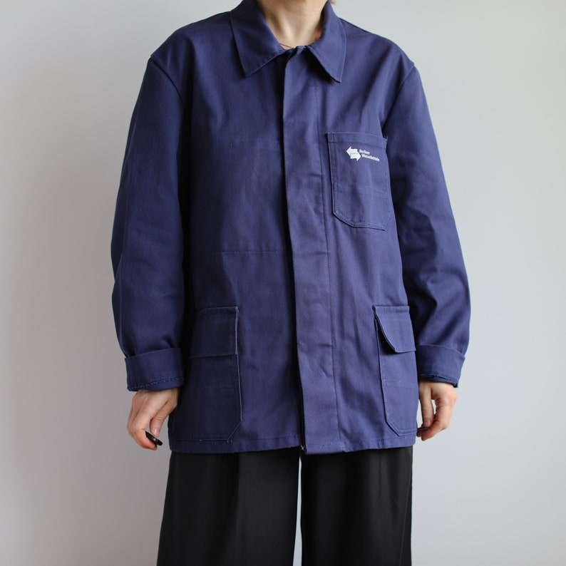 May include: A navy blue work jacket with a zipper closure and two large pockets on the front. The jacket has a white logo on the right chest pocket that says "Union des Transports Publics Genevois".