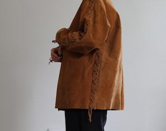 Puede incluir: Chaqueta de ante marrón con mangas largas y corte holgado. Las mangas y las costuras laterales presentan cordones decorativos. La chaqueta se combina con pantalones negros. El estilo general es vintage y bohemio.