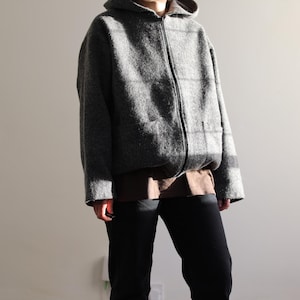 May include: A grey hooded jacket with a full-length zip and two front pockets. The jacket is made of a textured fabric and is worn over a brown top and black trousers. The jacket's hood is up, and the person is standing in front of a plain wall.