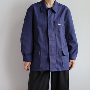 May include: A navy blue work jacket with a white logo that says "Atelier de Construction" on the left chest pocket. The jacket has two large pockets on the front and a button-up closure.