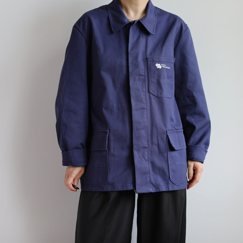May include: A navy blue work jacket with a white logo that says "Atelier de Construction" on the left chest pocket. The jacket has two large pockets on the front and a button-up closure.
