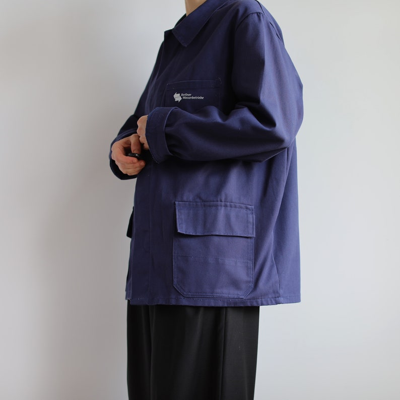 May include: A dark blue work jacket with a patch on the left chest that reads "S&uuml;dtiroler Wasserrettung". The jacket has a large pocket on the left side and a button-up closure.