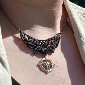 May include: A black and gold-toned necklace with a compass pendant. The necklace features a detailed wing-like design with intricate patterns. The pendant is silver-colored with a compass rose design. The necklace is worn over a black top and a tan jacket.