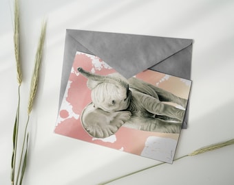 May include: A greeting card featuring a detailed illustration of a baby elephant in shades of gray, set against a pink and peach watercolor background. The card is partially covered by a gray envelope, with wheat stalks in the foreground.