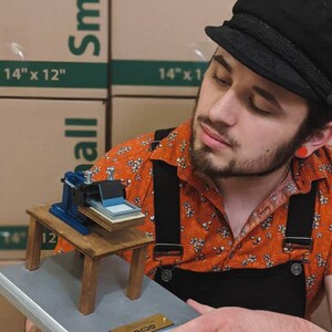 May include: A small wooden model of a screen printing press on a silver plaque with a gold plaque that says "BOD". The model is being held by a person wearing a black cap, an orange shirt with a floral pattern, and black overalls.