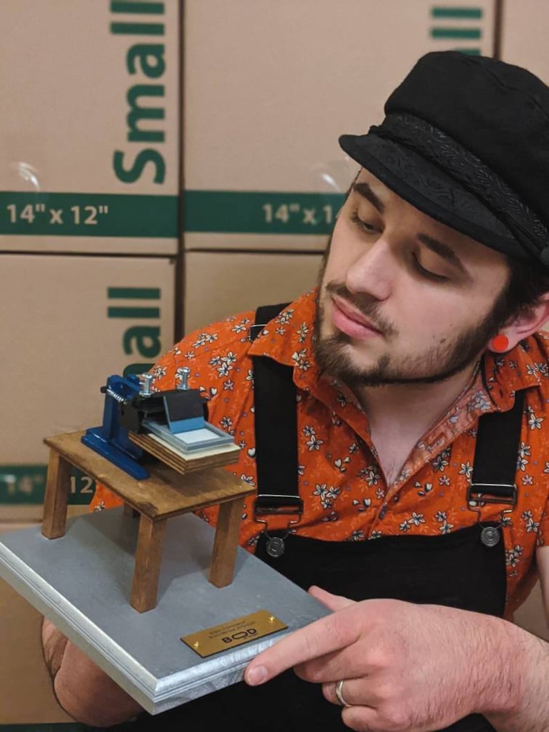 May include: A small wooden model of a screen printing press on a silver plaque with a gold plaque that says "BOD". The model is being held by a person wearing a black cap, an orange shirt with a floral pattern, and black overalls.