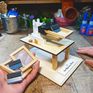May include: A small wooden table with a screen printing press and a sign that reads "The Spooky Shark". There are three screen printing frames on the table and a hand holding two more frames.