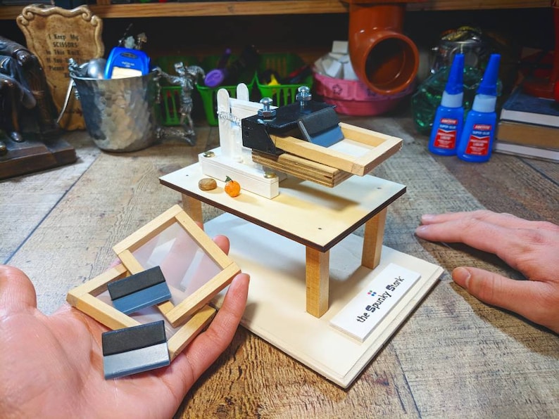 May include: A small wooden table with a screen printing press and a sign that reads "The Spooky Shark". There are three screen printing frames on the table and a hand holding two more frames.