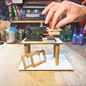 May include: A miniature wooden workbench with a small black clamp and two wooden frames. The workbench is on a white surface.