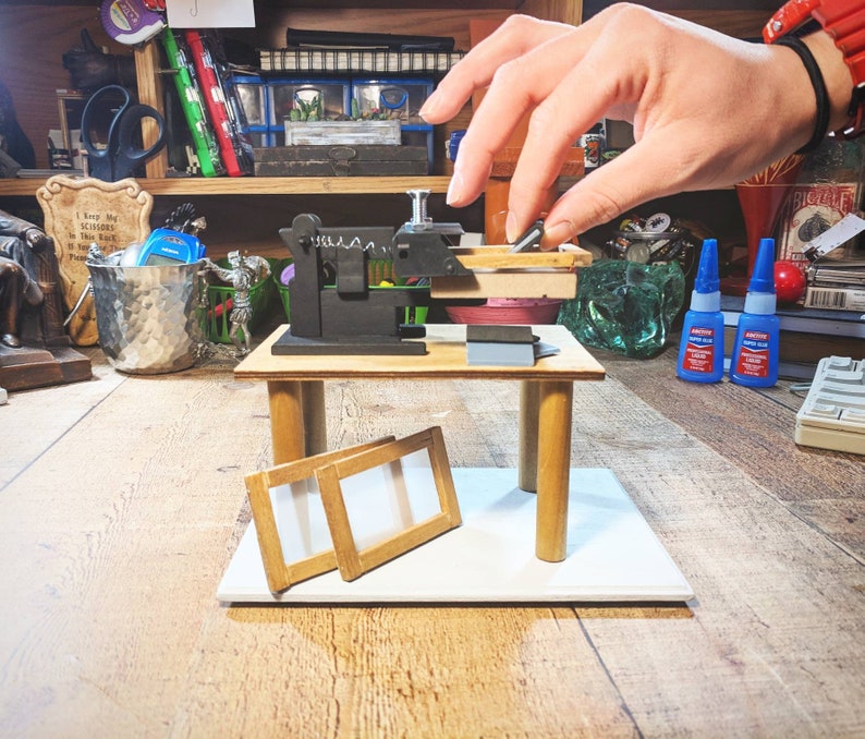 May include: A miniature wooden workbench with a small black clamp and two wooden frames. The workbench is on a white surface.