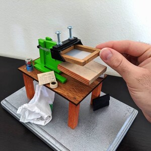 May include: A miniature green and black screen printing press with a wooden frame and a can of spray paint on a wooden table. The press is attached to a wooden base with a white cloth and a roll of tape.