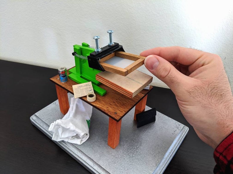 May include: A miniature green and black screen printing press with a wooden frame and a can of spray paint on a wooden table. The press is attached to a wooden base with a white cloth and a roll of tape.